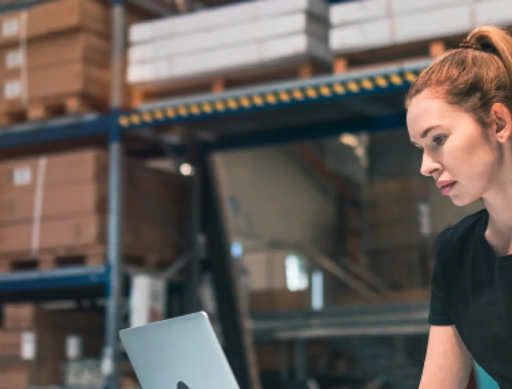 A woman with a ponytail is working on a laptop in a warehouse. She is wearing a black shirt and is surrounded by shelves filled with boxes.