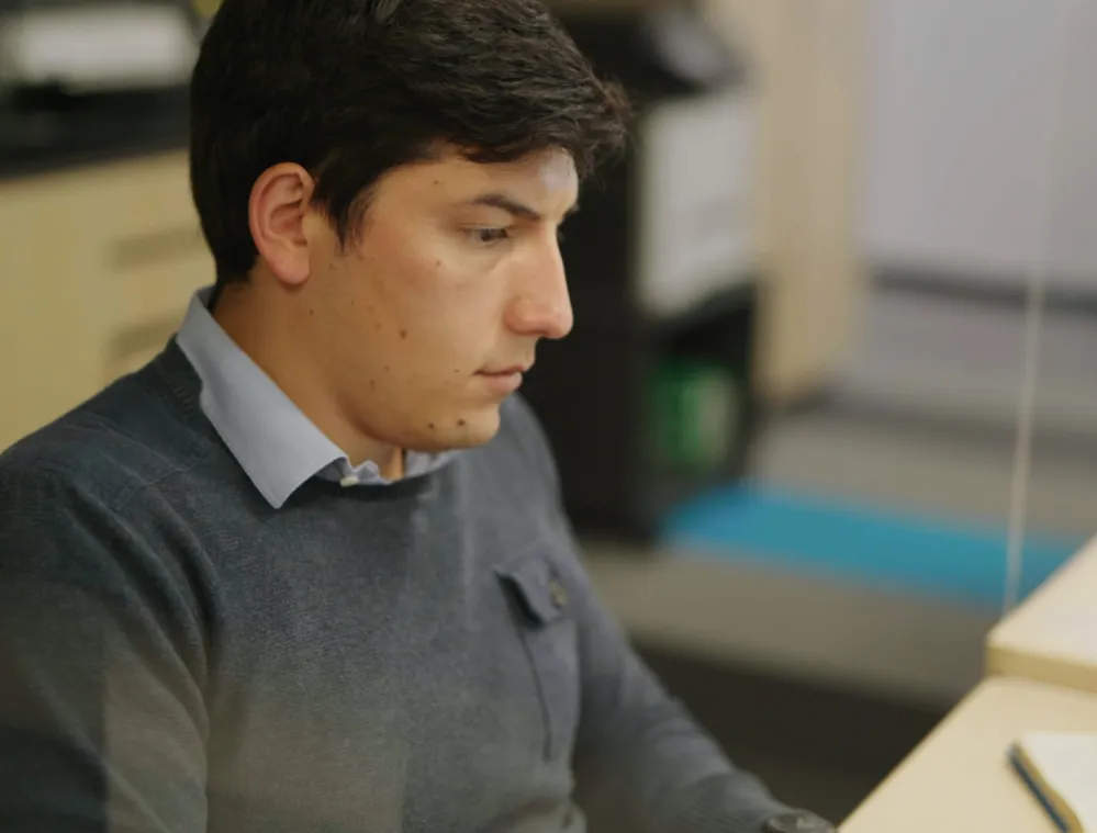 A person with short dark hair, wearing a gray sweater and light blue shirt, is focused on a task at a desk. A notebook and a smartphone are visible on the desk. The background is blurred, showing office furniture.