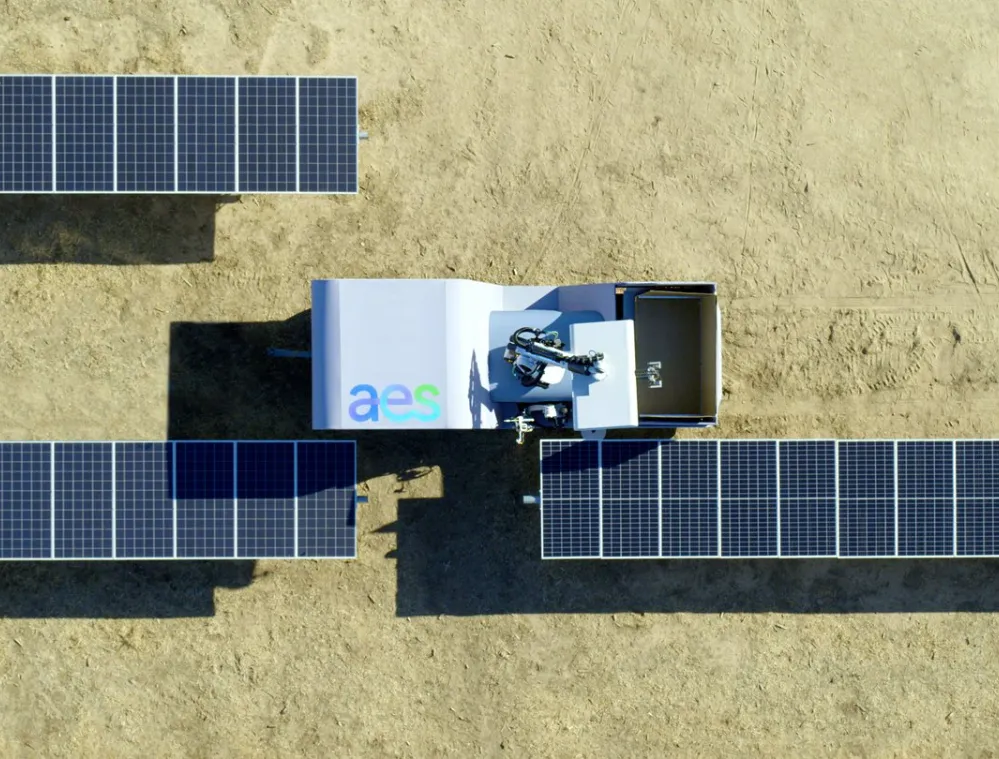 Aerial view of a solar panel installation on a dirt surface with a machine labeled 'aes' positioned between two rows of panels.