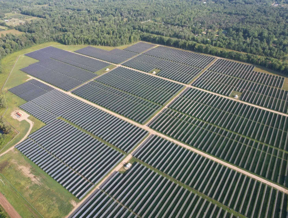 Aerial view of a large solar farm with rows of solar panels arranged in a grid pattern, surrounded by green forested areas.