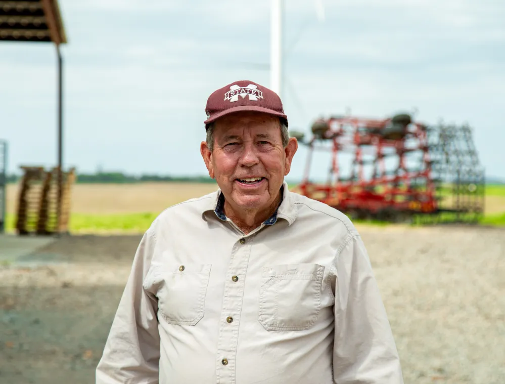 An older man wearing a beige shirt and maroon cap stands in front of a wind turbine and farm equipment on a cloudy day.