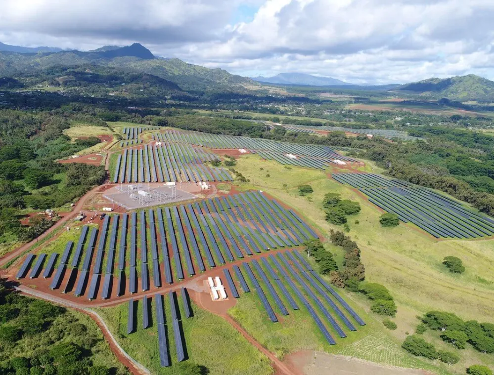 Aerial view of a large solar farm with rows of solar panels installed on green fields, surrounded by trees and mountains under a partly cloudy sky.
