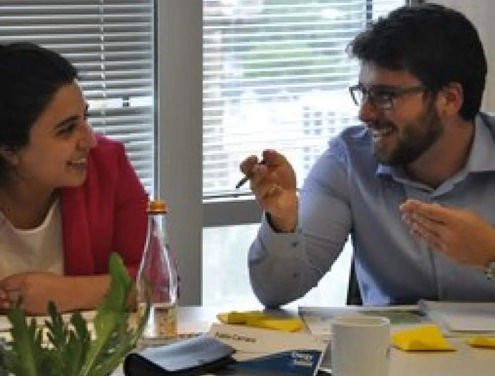 Two colleagues sit at a table in an office, smiling and having a friendly conversation. Papers, notebooks, a coffee mug, and a water bottle are on the table, with a window and blinds in the background.