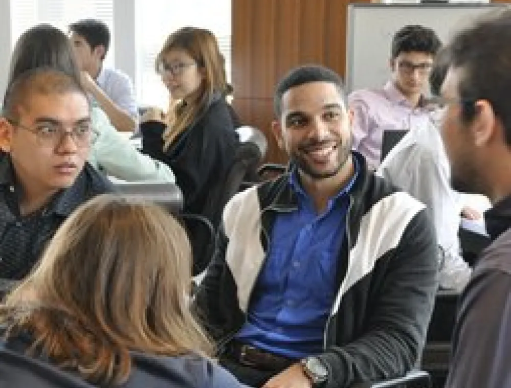 A group of young adults sitting in an office setting, engaged in conversation and group discussion. Some are smiling and interacting, while others are listening attentively. The background shows windows and a whiteboard.