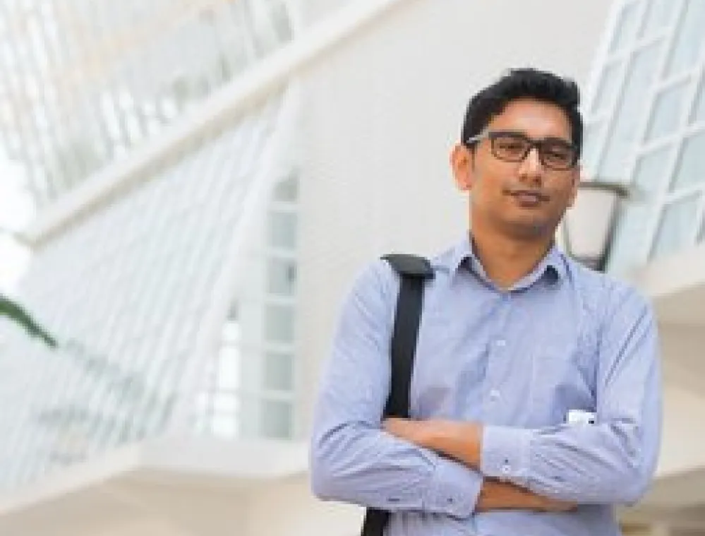 Man wearing glasses and a blue shirt stands with arms crossed in front of a modern glass building, carrying a shoulder bag and looking confidently at the camera.