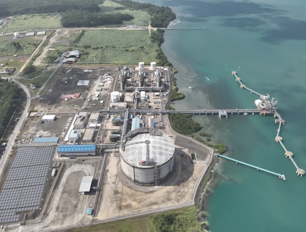 Aerial view of an industrial facility with large storage tanks, solar panels, and pipelines extending into the turquoise sea. The complex is surrounded by green fields and bordered by a coastline, with docks and infrastructure reaching into the water.