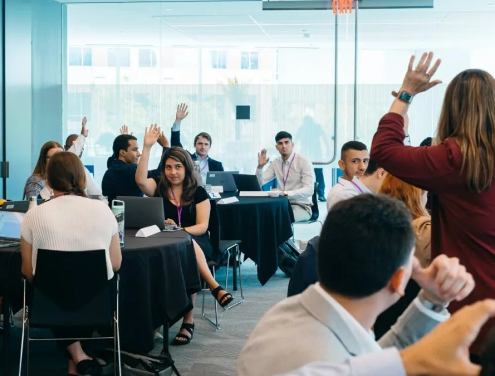 A group of adults in a modern conference room participate in a workshop, with several people raising their hands to answer a question. Laptops and notepads are on the tables, and a facilitator stands at the front engaging with the group.