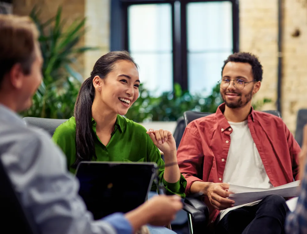 Young professionals participate in a collaborative discussion during a meeting in a modern office, with participants seated in a circle reviewing materials and sharing ideas.