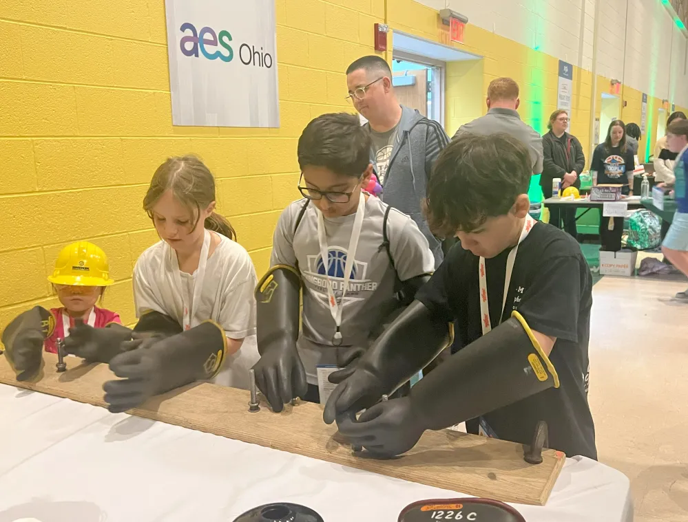 Children wearing oversized safety gloves performing a hands-on activity with a wooden board at an AES Ohio Safety 101 event.
