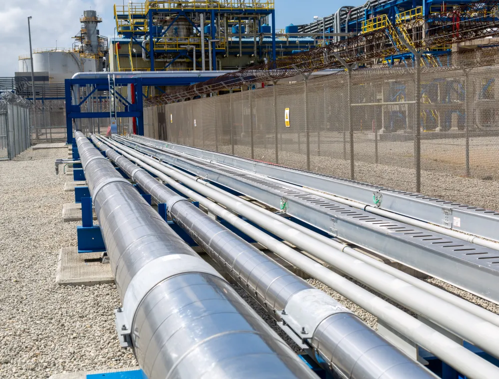 Industrial pipelines running alongside a wire mesh fence at a facility with large processing towers in the background.