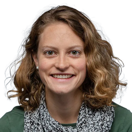 Portrait of a person with curly brown hair, smiling, wearing a green shirt and a patterned scarf, against a neutral background.