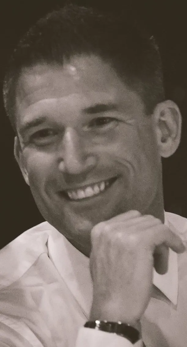Black and white photo of a smiling man with short hair, wearing a white shirt, resting his chin on his hand.