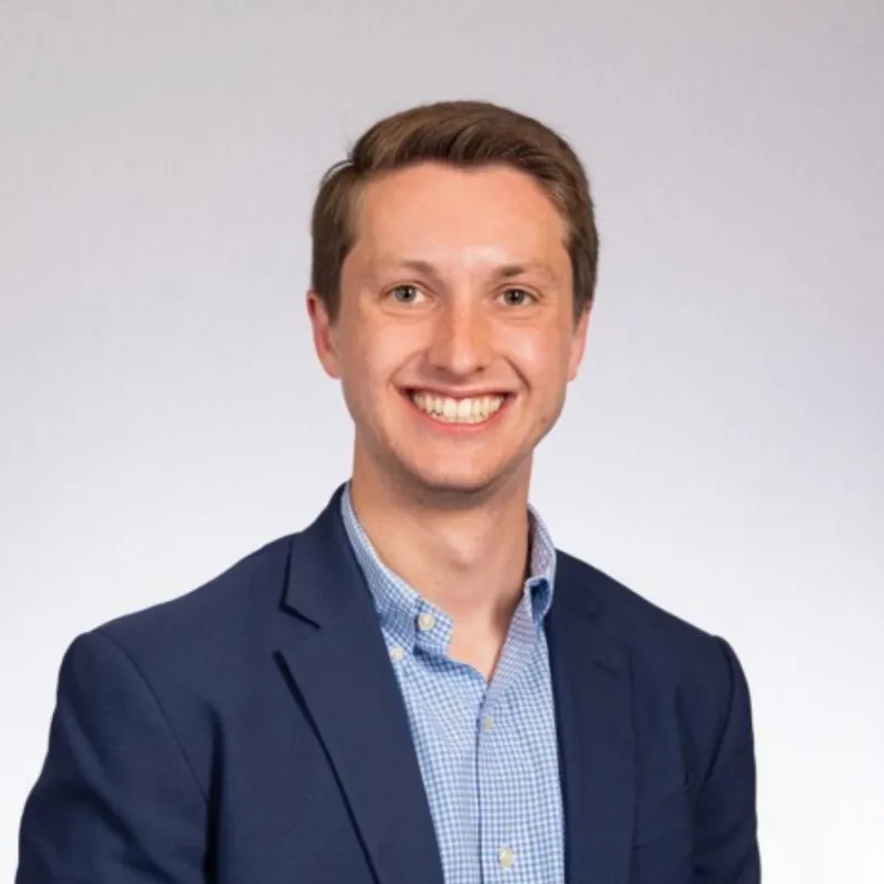 Smiling person wearing a blue blazer and checkered shirt against a light background.