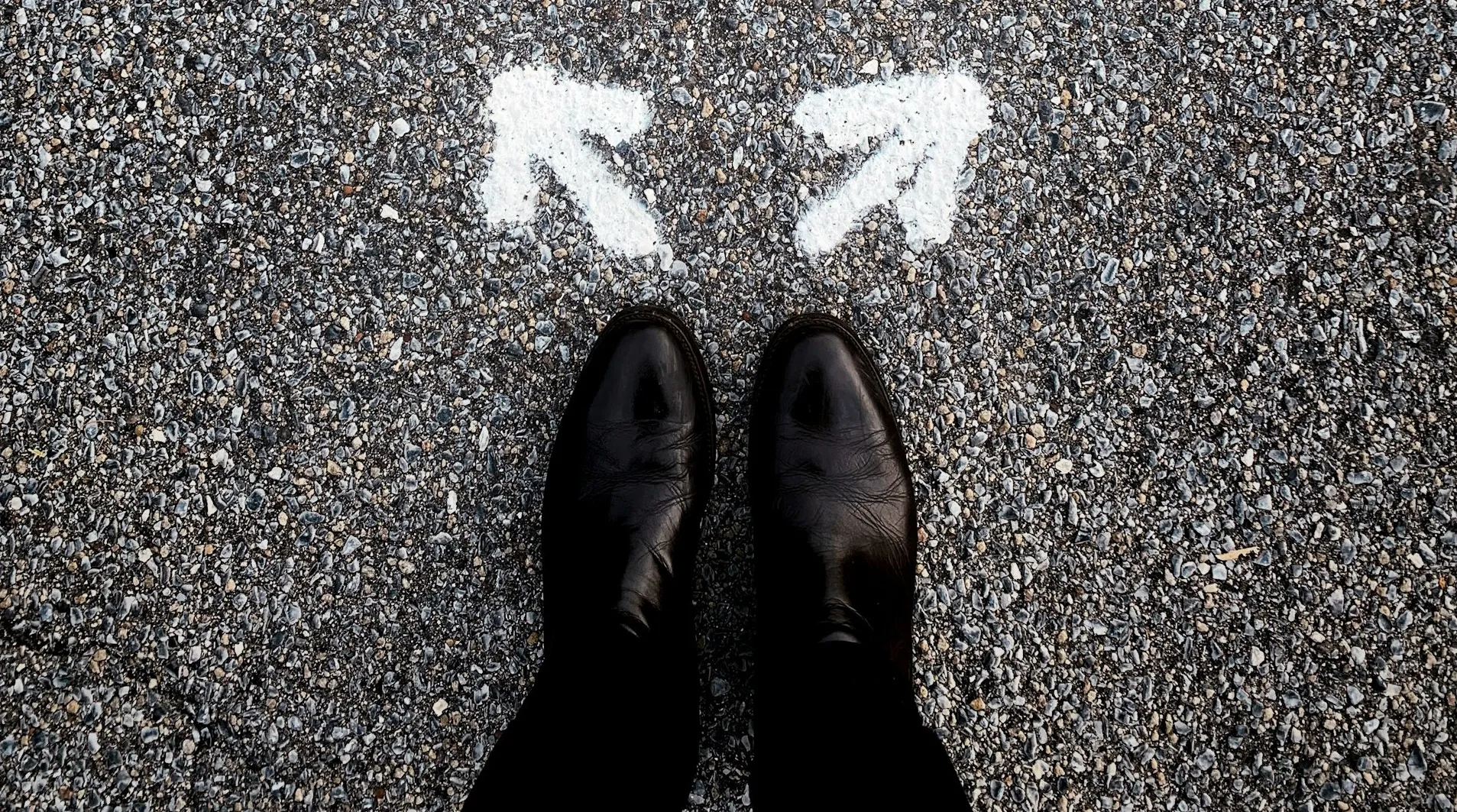 A person wearing black shoes stands on a gravel road in front of two white arrows painted on the ground, pointing in different directions, symbolizing a choice or decision.