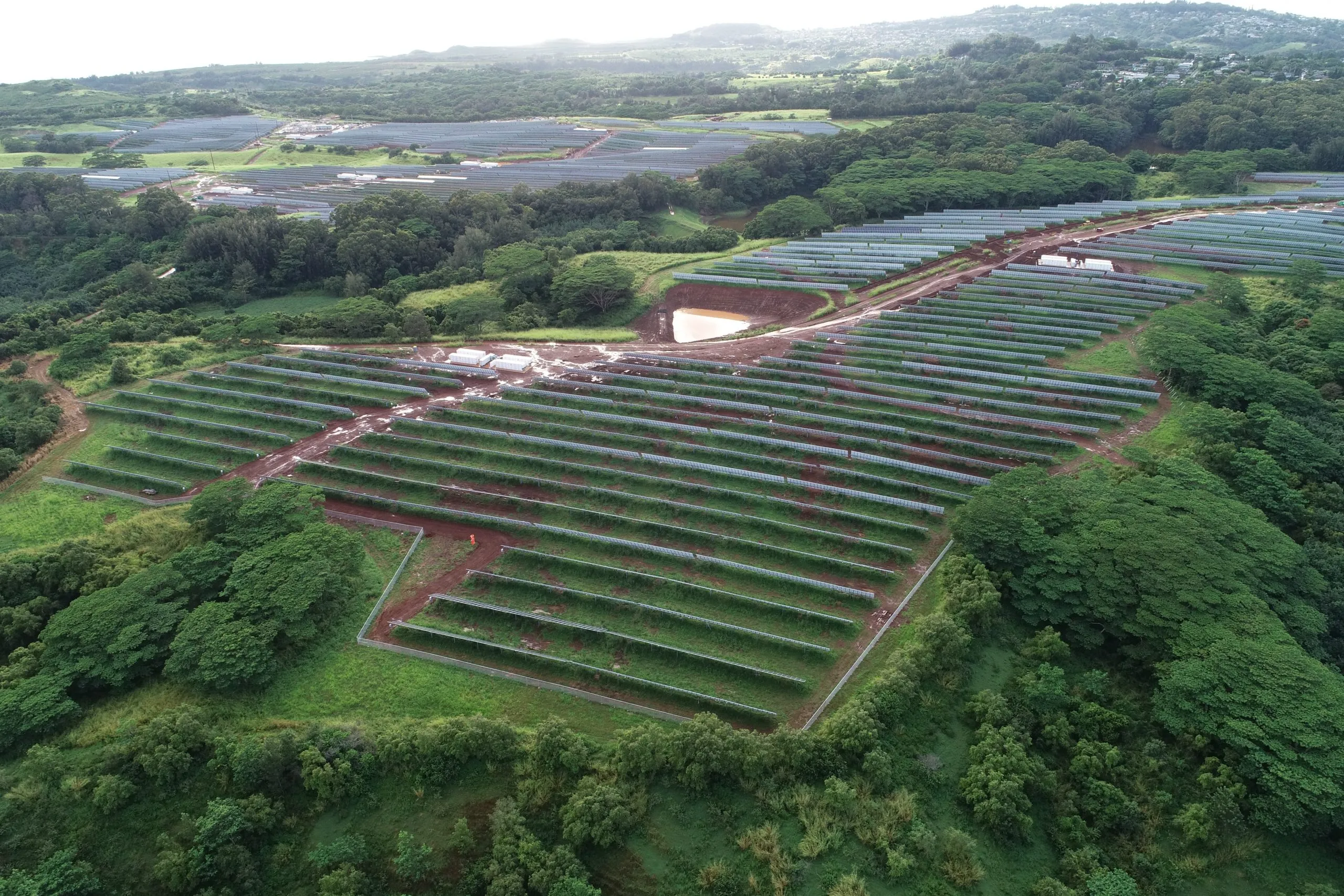 Aerial view of a large solar farm with rows of solar panels on a green landscape surrounded by trees and hills.