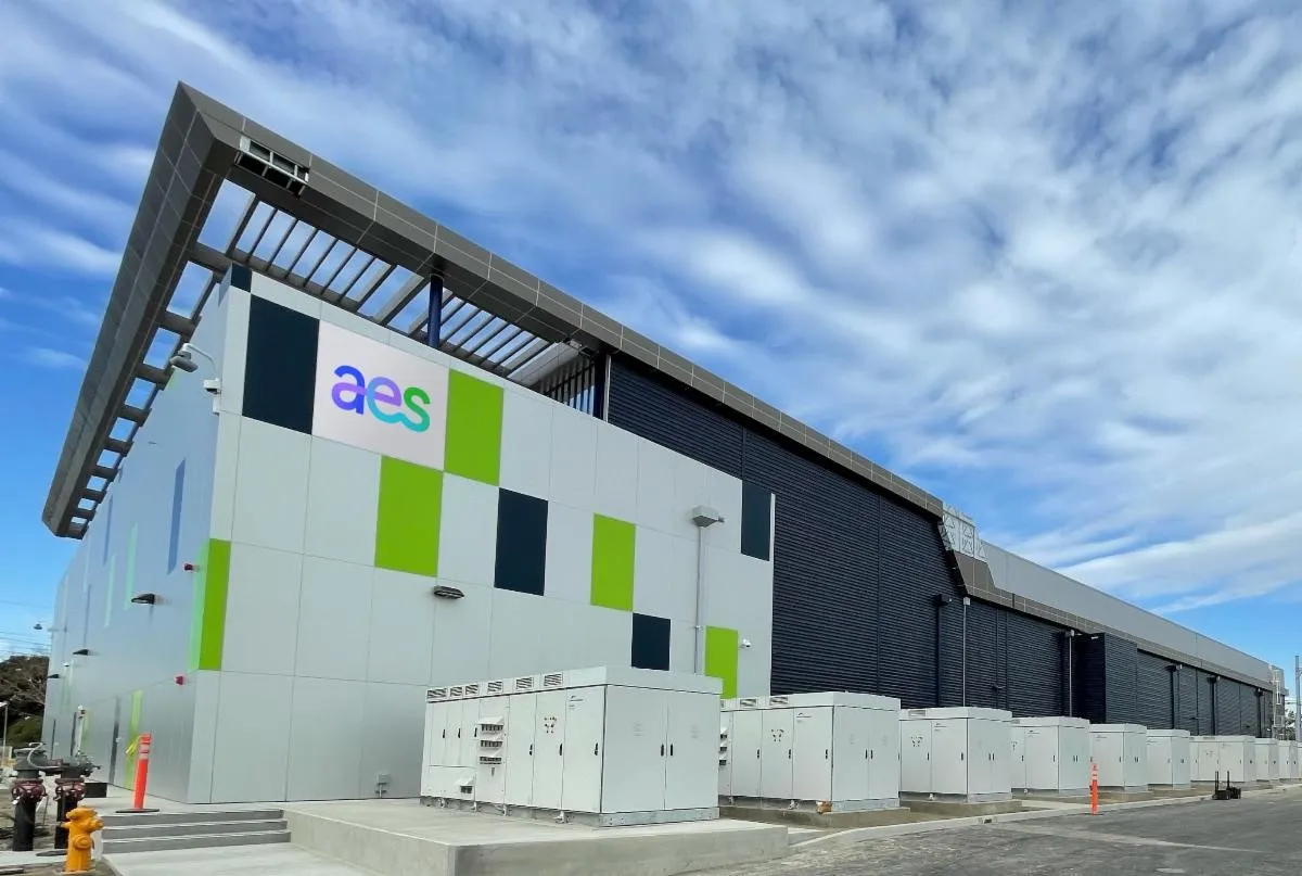 A modern industrial building with a geometric design and the letters 'aes' on the facade. The building features a mix of white, green, and dark panels, with several electrical units in front. The sky is partly cloudy.