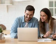 A man and woman smiling while looking at a laptop in a kitchen. The man is wearing glasses and a denim shirt, and the woman is holding a pen. A notebook and calculator are on the table.