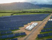 Aerial view of a large solar farm with numerous solar panels arranged in rows, set against a backdrop of green fields and distant mountains under a cloudy sky.