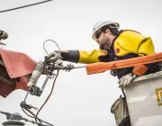 A utility worker wearing safety gear, including a hard hat, sunglasses, and insulated gloves, operates from a bucket lift.