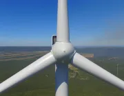 Aerial view of a wind turbine's hub and blades against a scenic landscape with fields and distant hills under a clear blue sky.