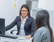 A woman in a black blazer and glasses is sitting at a desk, smiling at another woman with long hair who is sitting across from her. 